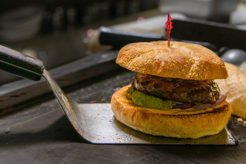 A gourmet burger with a patty, green sauce, and red onions on a spatula.