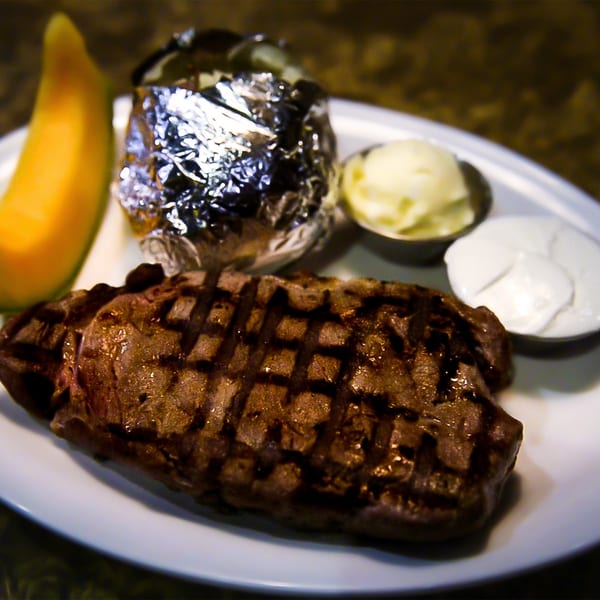 Grilled steak with a foil-wrapped baked potato, butter, sour cream, and a melon slice.