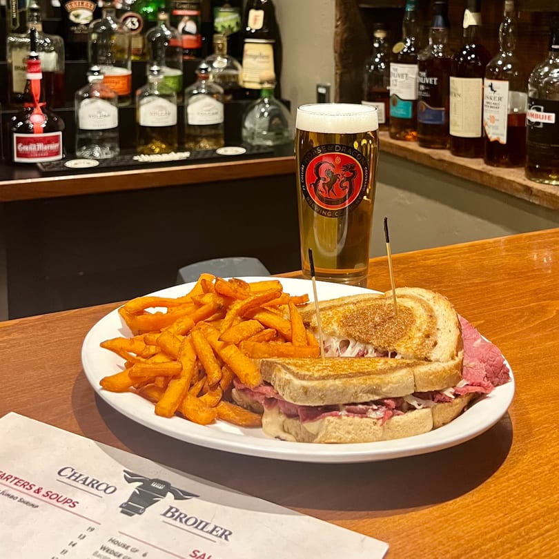 Reuben sandwich, sweet potato fries, and a Horse & Dragon beer on a wooden bar with liquor bottles.